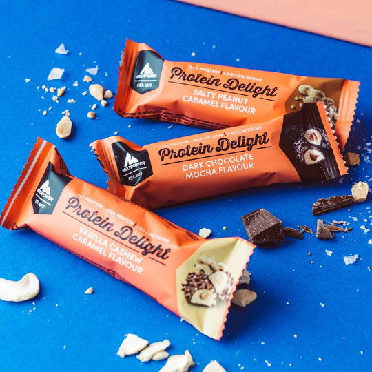 Alle Sorten des Multipower Protein Delight Riegels auf einem Tisch arrangiert – daneben Riegelstücke als Einblick in Textur und Geschmack.