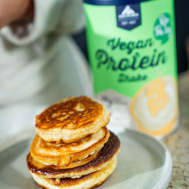 Multipower Vegan Protein Shake 420g im Hintergrund – im Vordergrund ein Teller mit frisch zubereiteten Pancakes als pflanzliche Genuss-Inspiration.
